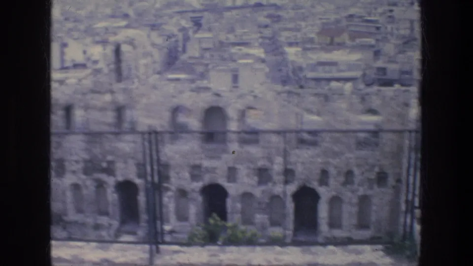 Athens ruins with arched openings and a metal barrier in front, with modern buildings filling the background.