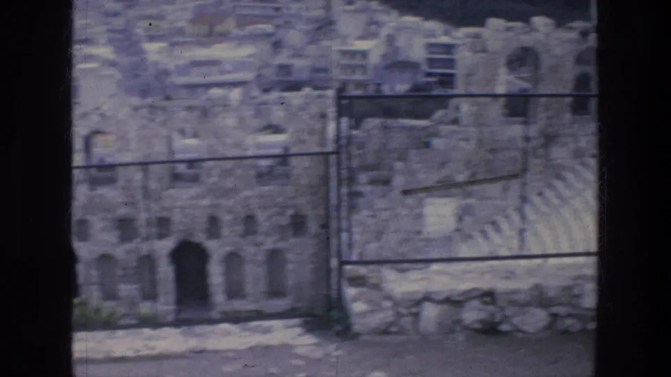 1976 Athens home-movie frame showing a weathered ruin, arched doorway, tiered seating, and metal fence in soft 8mm color.