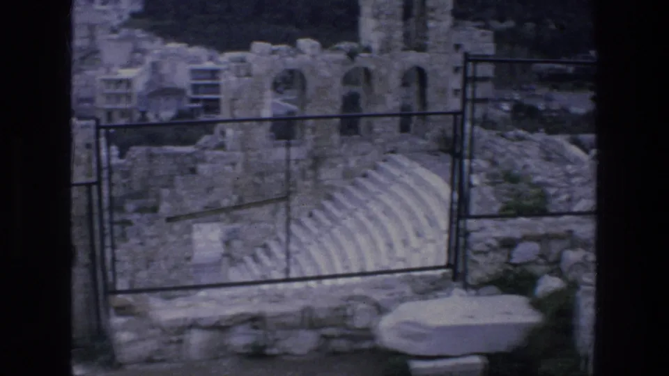 Fenced view of ancient-looking stone seating and ruined walls in Athens, with rubble and dark film edges visible.