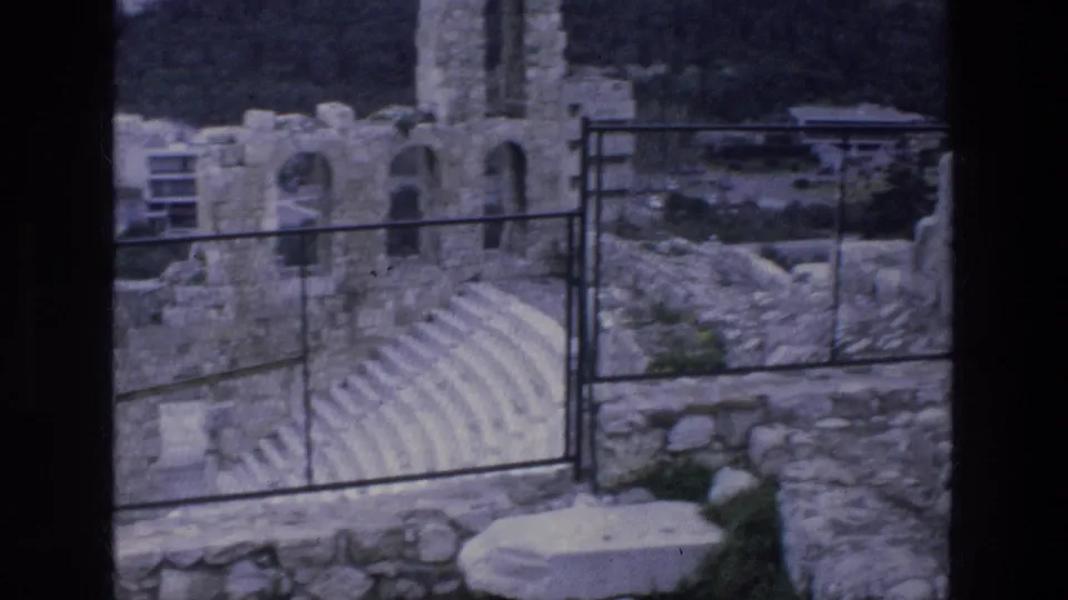 1976 Athens home-movie view of tiered stone seating, arched ruins, rocky ground, and a metal site railing.