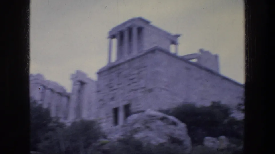 1976 Athens home-movie frame showing a stone ruin with small columns, wall openings, rocks, and low greenery.