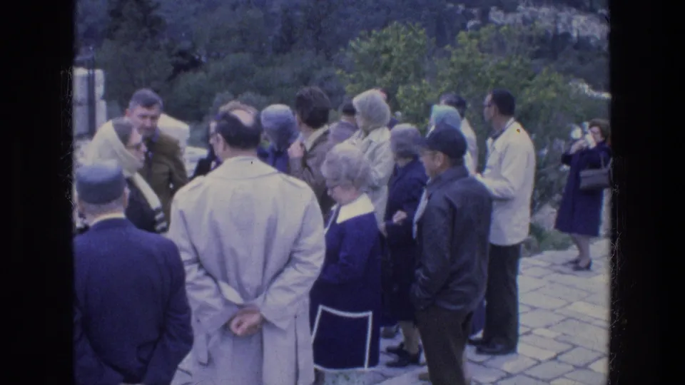 Adult tourists at an Athens archaeological site stand in a loose semicircle, with stone paving, greenery, and ruins nearby.