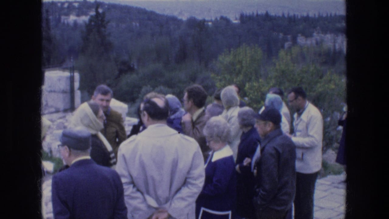 1976 Athens home-movie frame showing a group of tourists in coats gathered beside stonework with hills beyond.