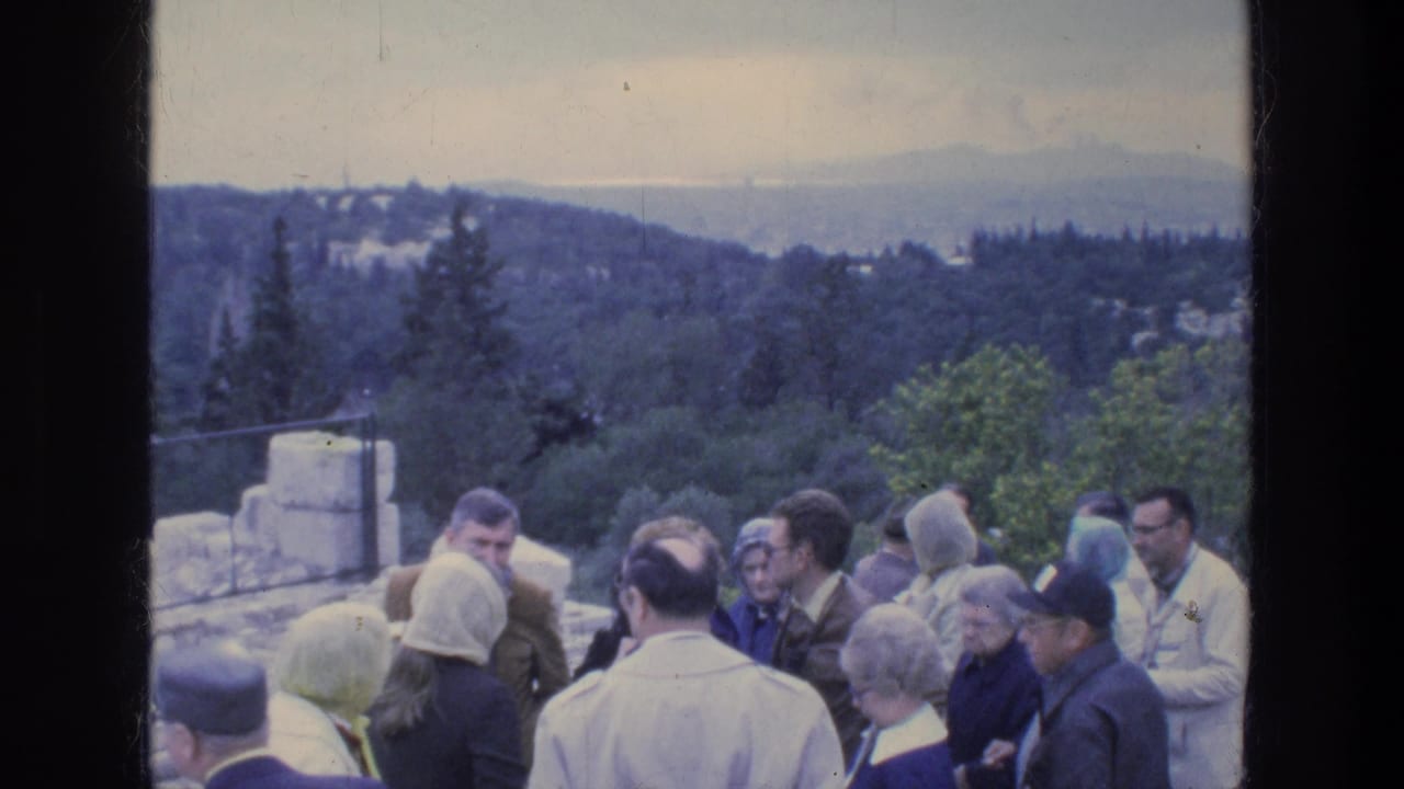 Tourists at an outdoor Athens site stand near ancient-looking masonry while the landscape opens behind them.