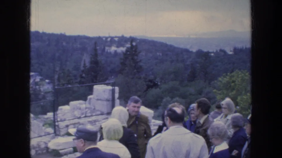 1970s home-movie view of tourists in coats on a stone-paved overlook with wooded hills behind them.