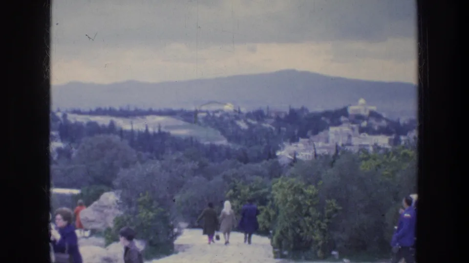 Athens hillside view with distant buildings and mountains, with visitors walking or standing near stone steps.