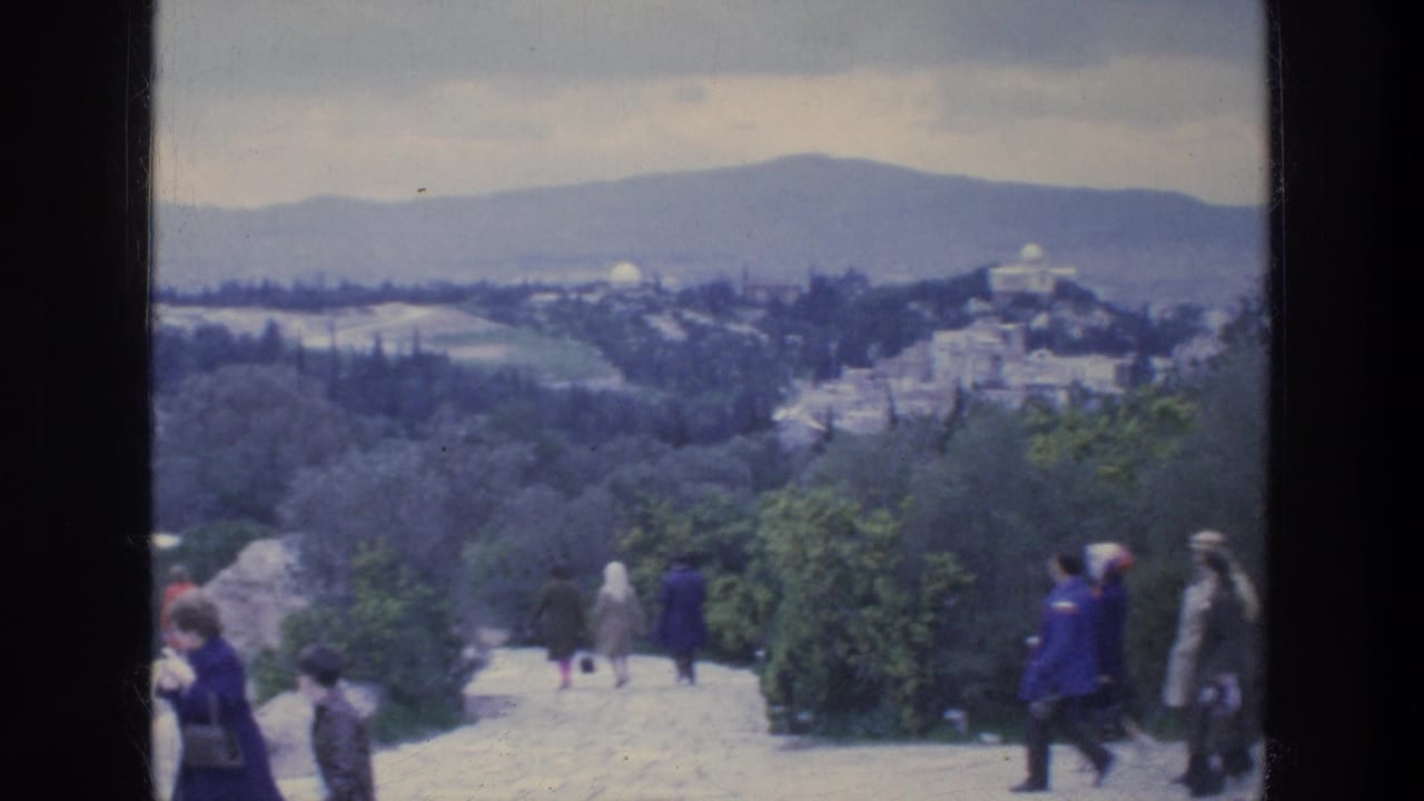 1976 Athens home-movie frame showing tourists on a stone overlook path with greenery, distant city buildings, and hills under a cloudy sky.