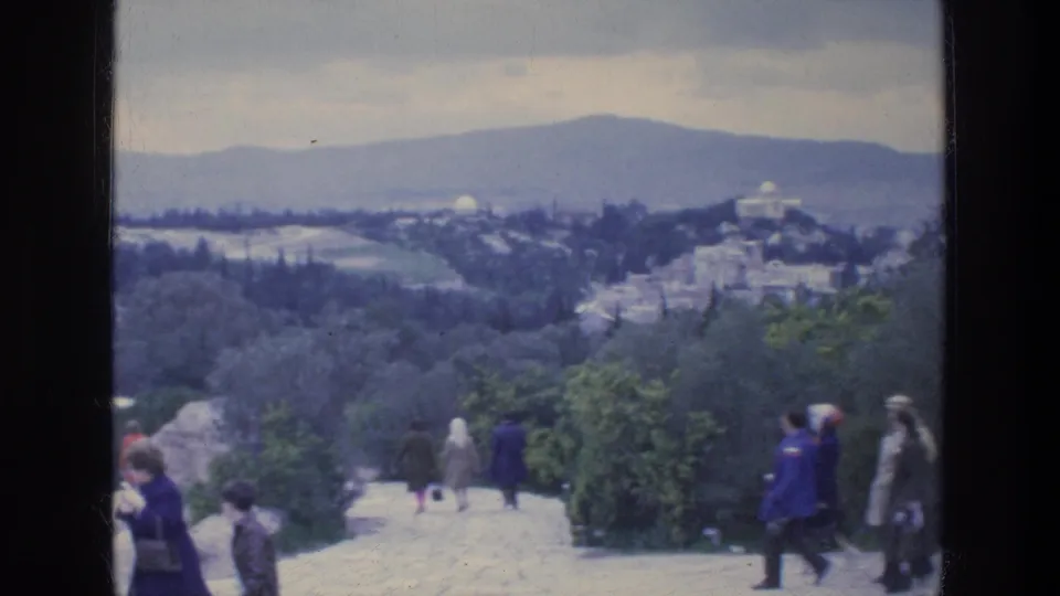 1976 Athens home-movie frame showing tourists on a stone overlook path with greenery, distant city buildings, and hills under a cloudy sky.