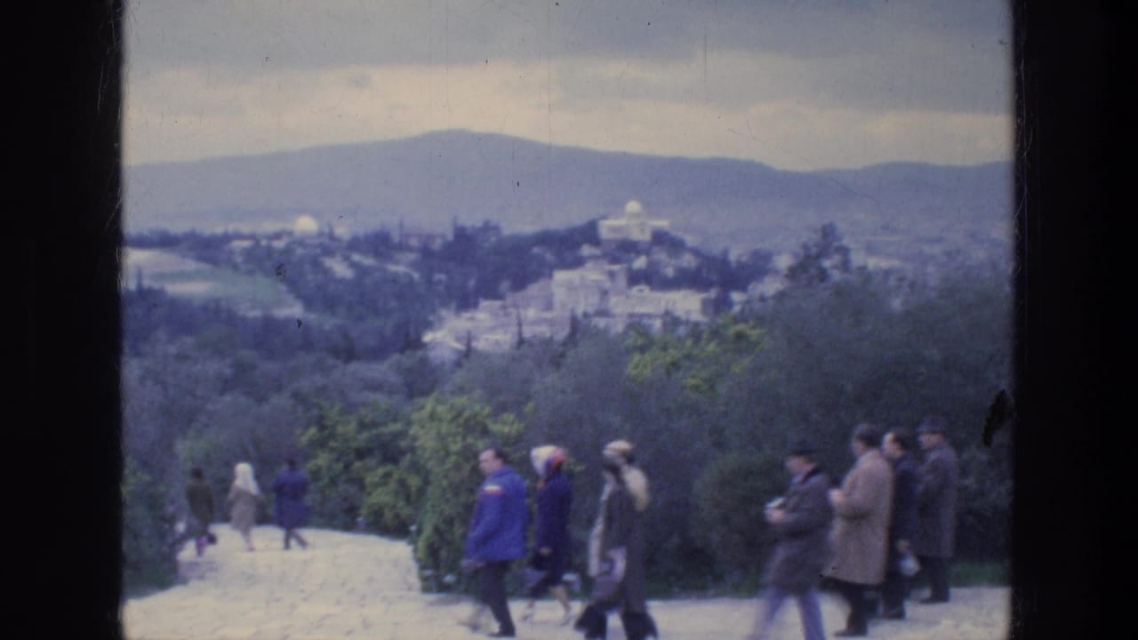 Groups of tourists walk across a hillside overlook with a city and hills beyond.