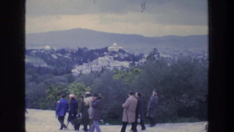 1976 Athens home-movie view of tourists in coats on a scenic path, with dark trees, city structures, and hazy hills beyond.
