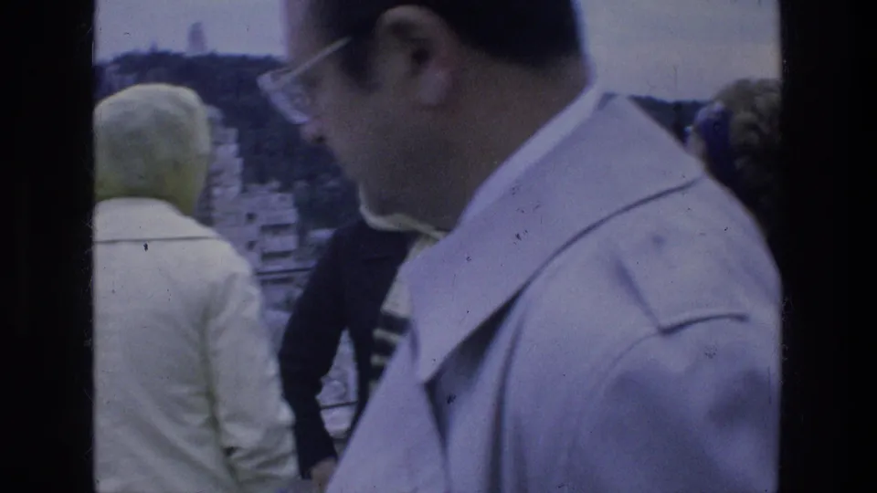Close 1976 Athens frame showing a man in glasses in the foreground, with other tourists and the hillside overlook behind him.