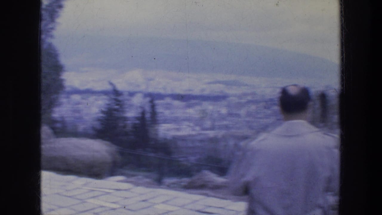 Man in a light jacket facing away toward a blurred city overlook and distant hills.