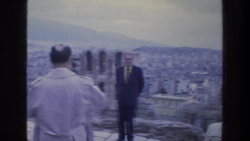 1970s Athens overlook scene with two adult men positioned against a broad city panorama and distant mountain slope.