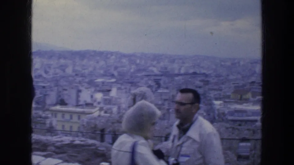Adult tourists at an Athens overlook, including a light-haired woman and a man in sunglasses, with the city panorama behind them.