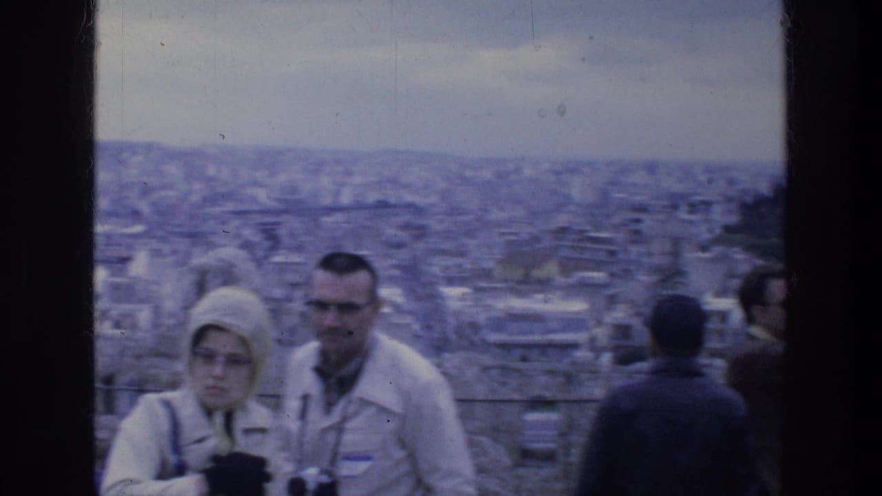 1976 Athens home-movie frame showing tourists in coats with cameras near a city overlook by ancient ruins.