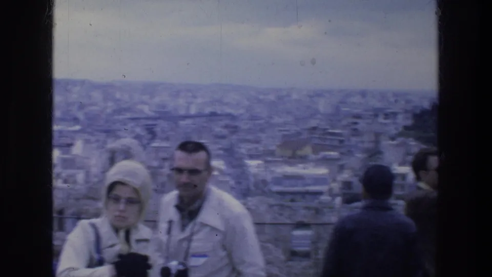 1976 Athens home-movie frame showing tourists in coats with cameras near a city overlook by ancient ruins.