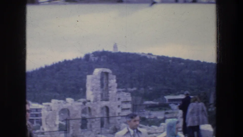 1976 Athens view of ancient ruins, railings, tourists and a wooded hillside above the surrounding city.