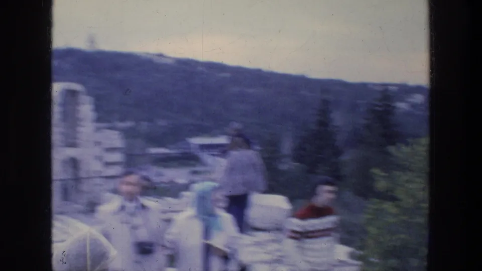 Handheld 8mm frame of tourists moving at an Athens ruin overlook, with hillside and urban background visible.
