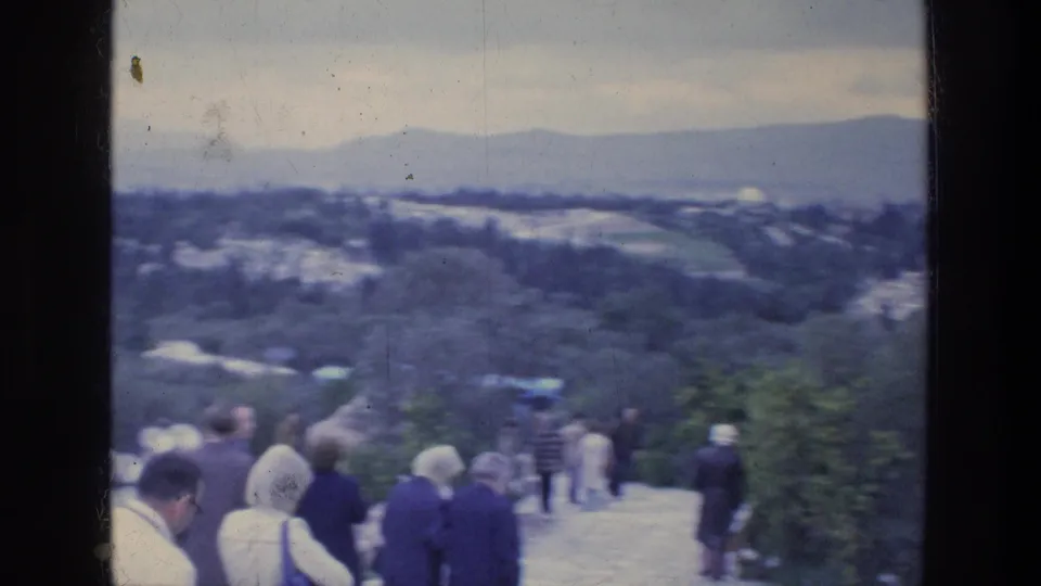 1976 Athens 8mm frame showing tourists walking down a scenic hill path with hazy hills and buildings beyond.
