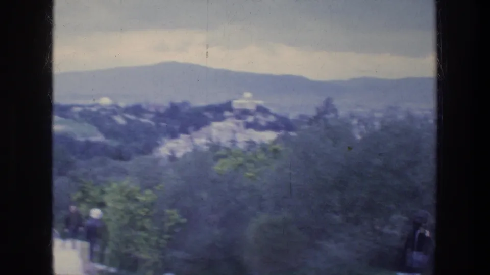 1976 Athens hilltop view across trees toward distant buildings and mountains, with tourists partly visible below.
