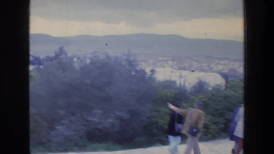 People in 1970s casual clothing walk along an Athens overlook path, one appearing to gesture toward the distant city.