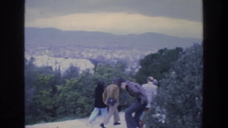 Several visitors in 1976 Athens walk along a park path, partly turned toward the view over trees and the distant city.