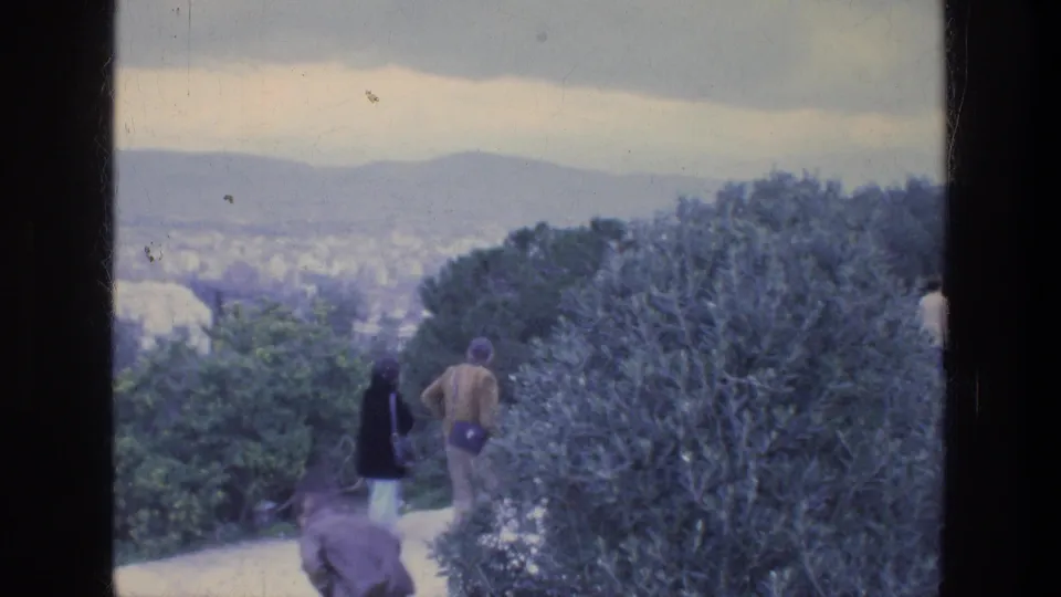 Final frame of a 1976 Athens overlook walk, with visitors on the path, a large shrub, and a hazy city-and-hill backdrop.