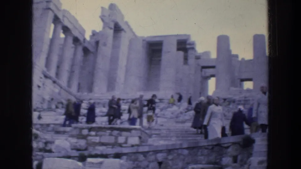 Wide 8mm frame of tourists crossing the Acropolis steps in Athens, with broken columns and stone platforms visible.