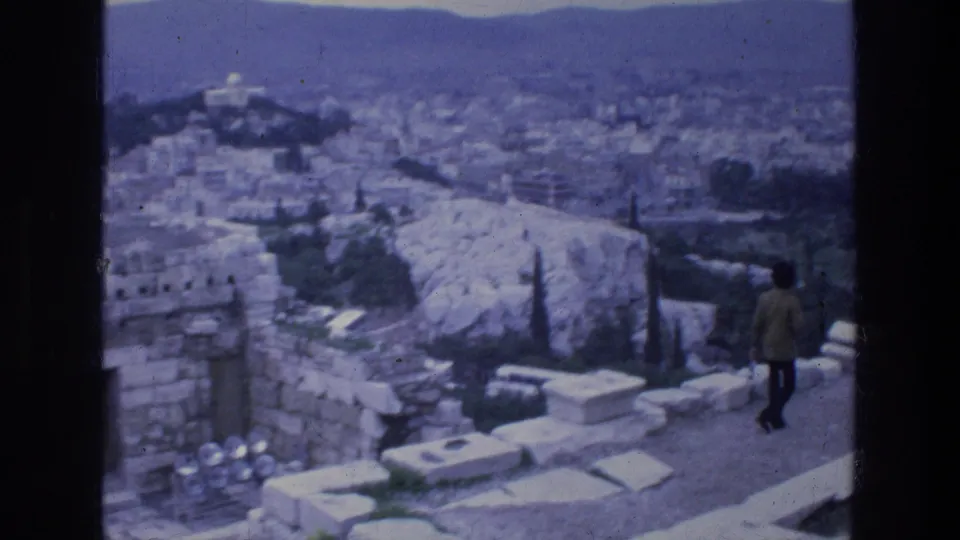 1976 8mm view from ancient ruins in Athens, with an adult visitor near stone blocks and the city spread below.