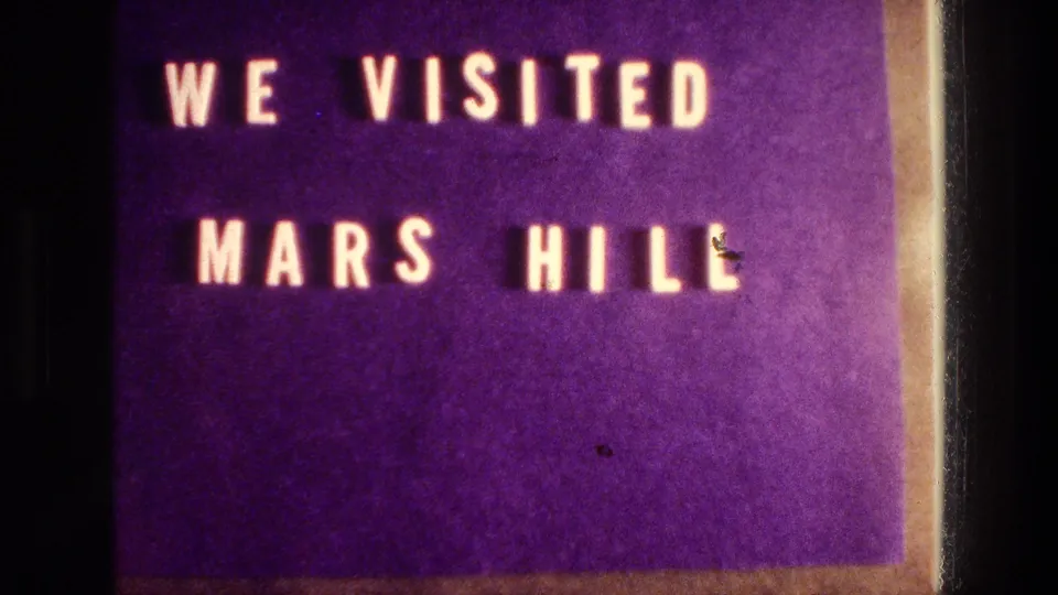 Close 8mm frame of the WE VISITED MARS HILL title card, with purple color, soft lettering, and visible dust specks.