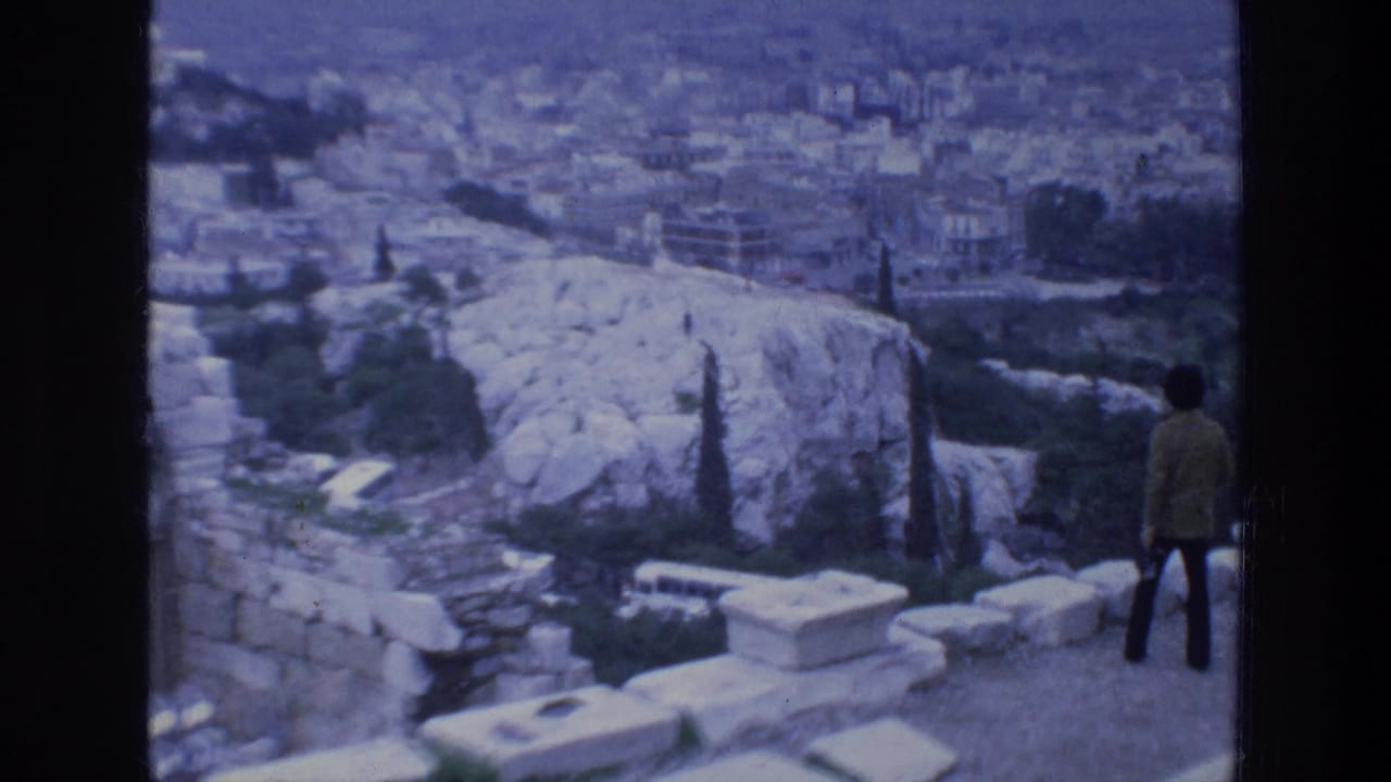 Tourist stands near stone ruins overlooking a rocky hill and city buildings.