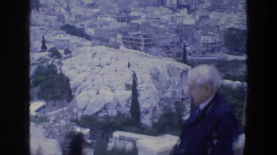 Older tourist moves through the foreground during a 1970s Athens Acropolis view over pale rock and dense city buildings.