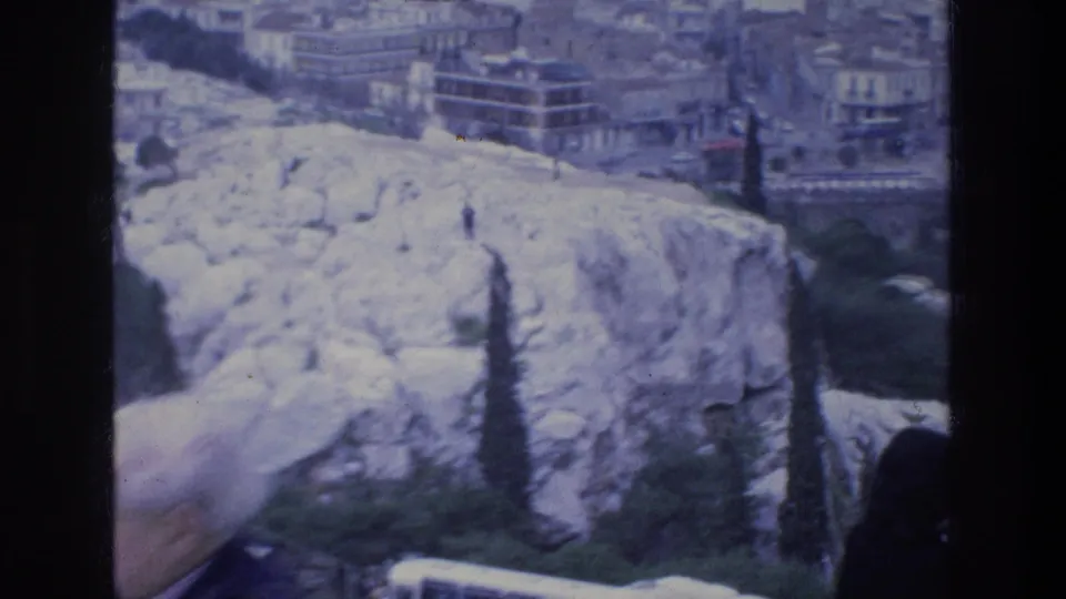 Acropolis overlook in Athens with pale rock, stone edges, trees, city buildings, and a blurred foreground visitor.