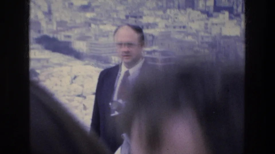 A suited man with glasses stands near pale Acropolis-area rocks, holding camera gear in 1976 Athens travel footage.
