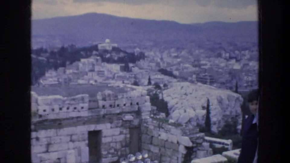 Wide 1976 8mm view over Athens from stone ruins, with pale rocky hillside, buildings, trees, and distant hills.
