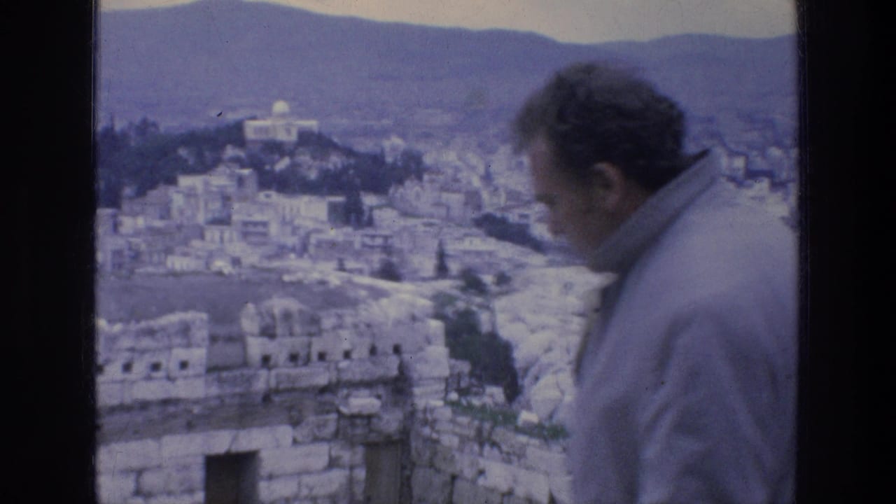 Man in a light jacket stands before stone ruins with Athens buildings and mountains beyond.