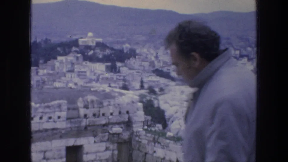 1976 8mm Athens view with an adult visitor in the foreground, ancient stonework below, city buildings, a white dome, and distant mountains.