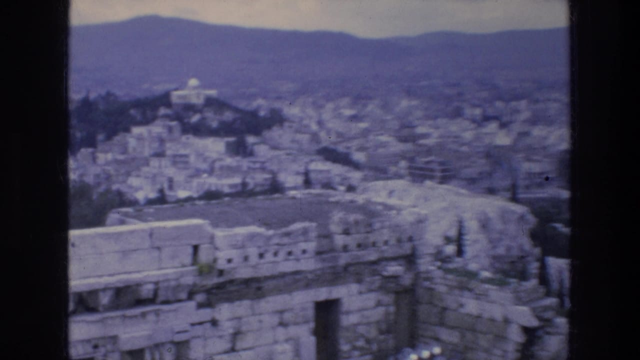 Stone ruins overlook a dense cityscape with a white domed building and mountains in the distance.