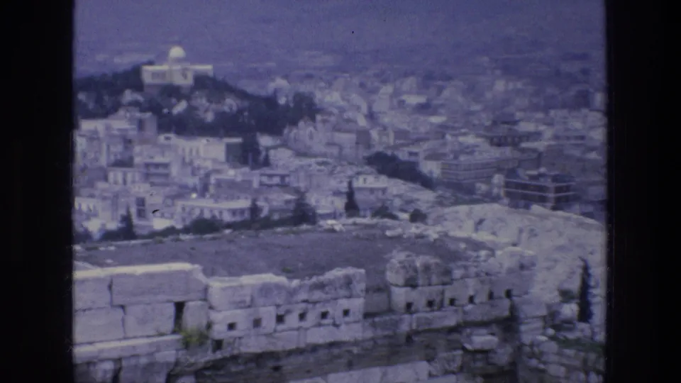 1976 8mm Athens overlook with ancient stone blocks in front, city blocks below, a white domed structure on a hill, and distant mountains.