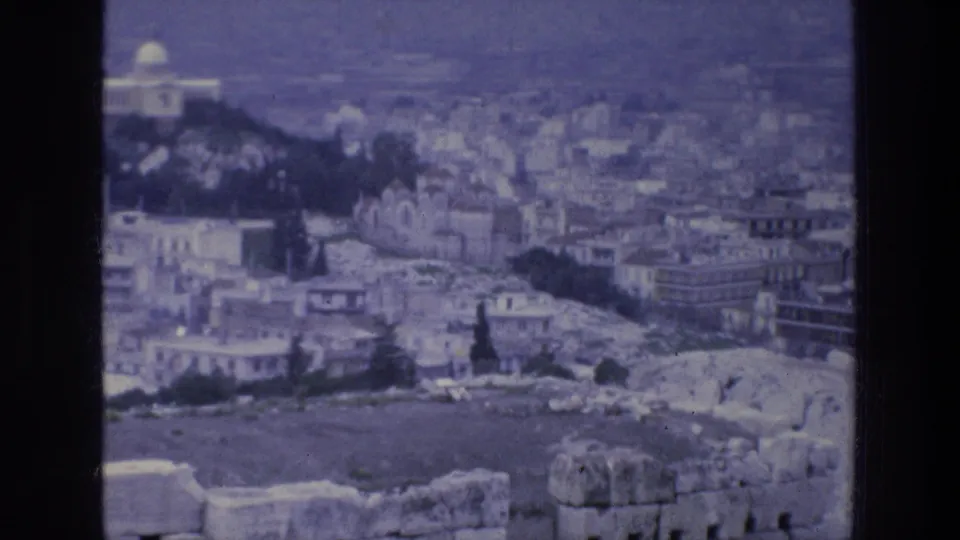 1976 8mm travel frame looking across Athens from an elevated ruin site, with stonework, city buildings, trees, a white dome, and mountains.