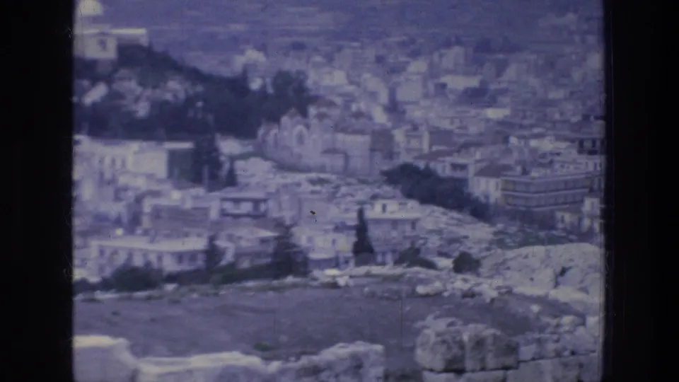 1976 8mm view over Athens buildings, rocky hillside, trees, and pale ruin-like stonework in soft blue-tinted focus.
