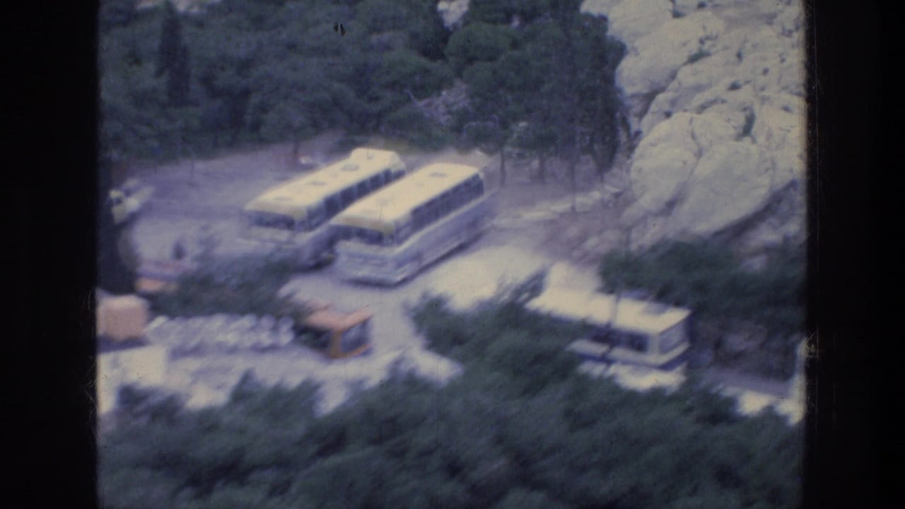 Parked tour buses beside trees and rocks in a hillside parking area.