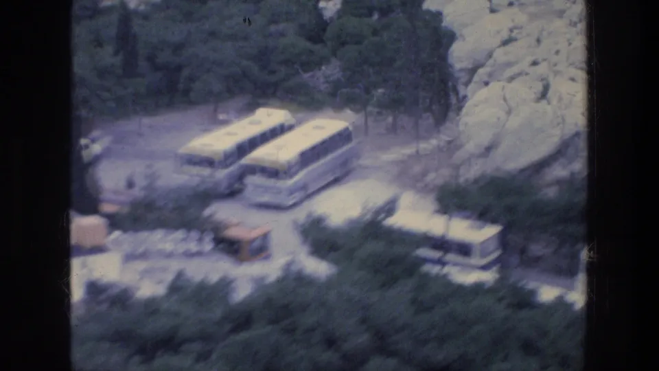 Blue-tinted 1976 Athens travel frame showing several parked tour buses near trees, rocky slopes, and a small roadside structure.