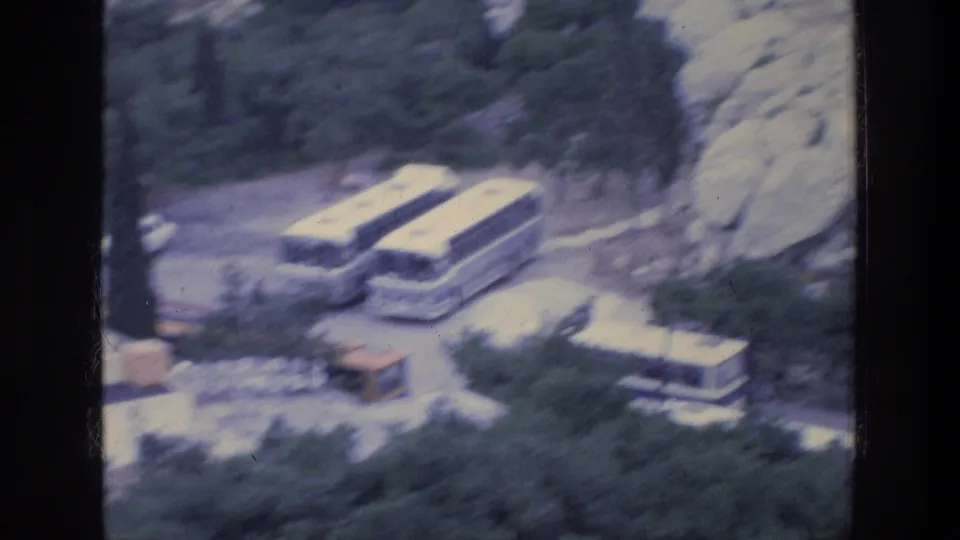 1976 Athens home-movie frame with parked tour buses, a small structure, trees, rocks, and a pale vehicle partly visible below.