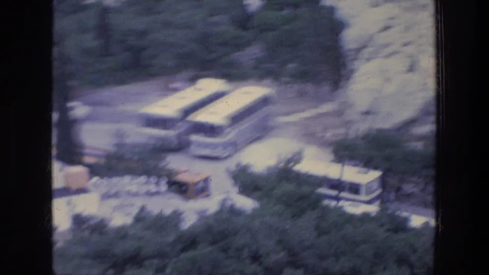 Late clip frame from 1976 Athens showing parked buses, a pale vehicle, trees, rocky hillside edges, and strong blue 8mm color shift.
