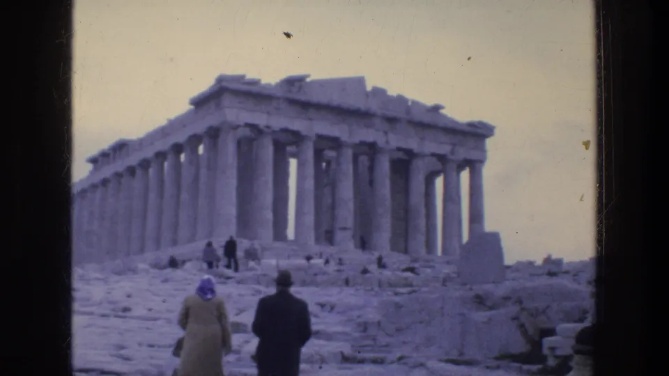1976 8mm travel frame showing tourists approaching the Parthenon in Athens, with columns, scattered ruins, and pale sky visible.