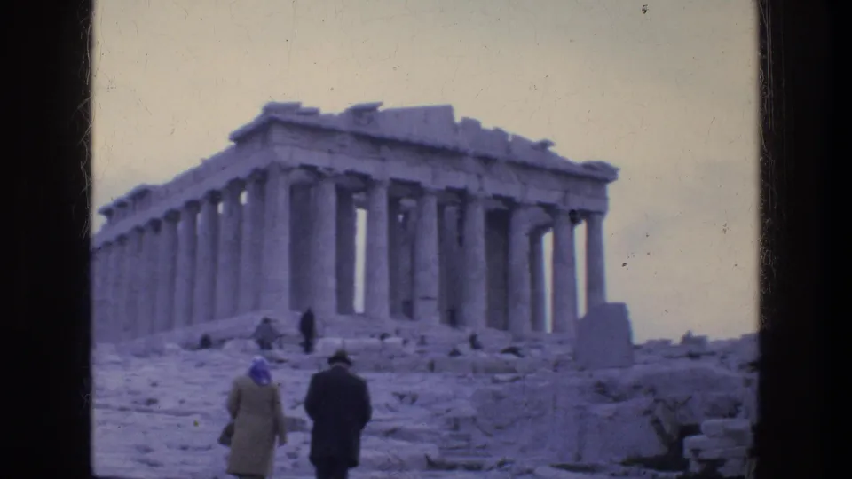 Athens travel footage frame with two visitors in period coats walking toward the Parthenon, seen through soft 8mm color grain.