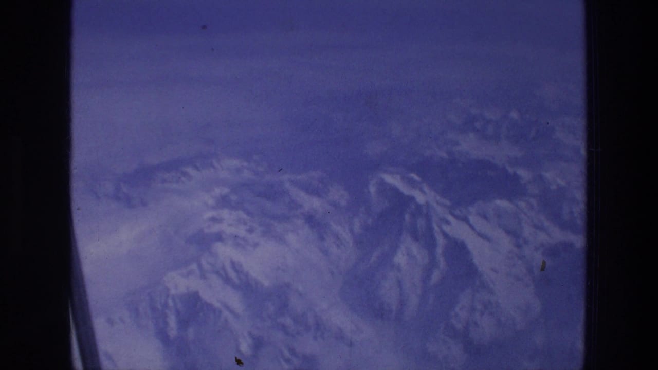 1976 8mm travel footage showing snowy mountain peaks below an airplane window, with blue-purple film color and grain.