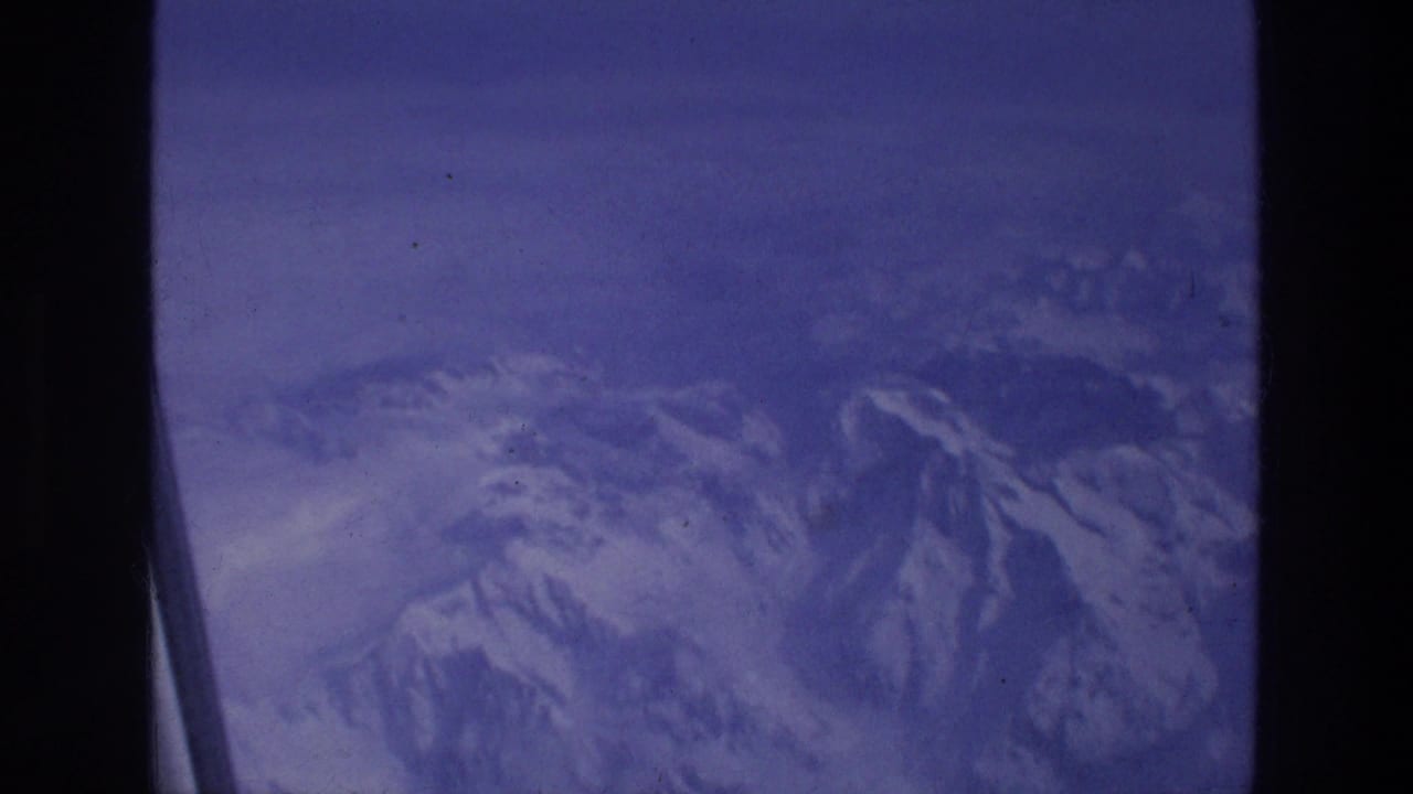 Aerial mountain view from a plane, with snow on the ridges and visible 8mm film grain from the 1976 Greece travel reel.