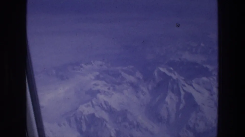 Snow-dusted mountain terrain below an airplane window, rendered in soft blue-purple 8mm home-movie color.